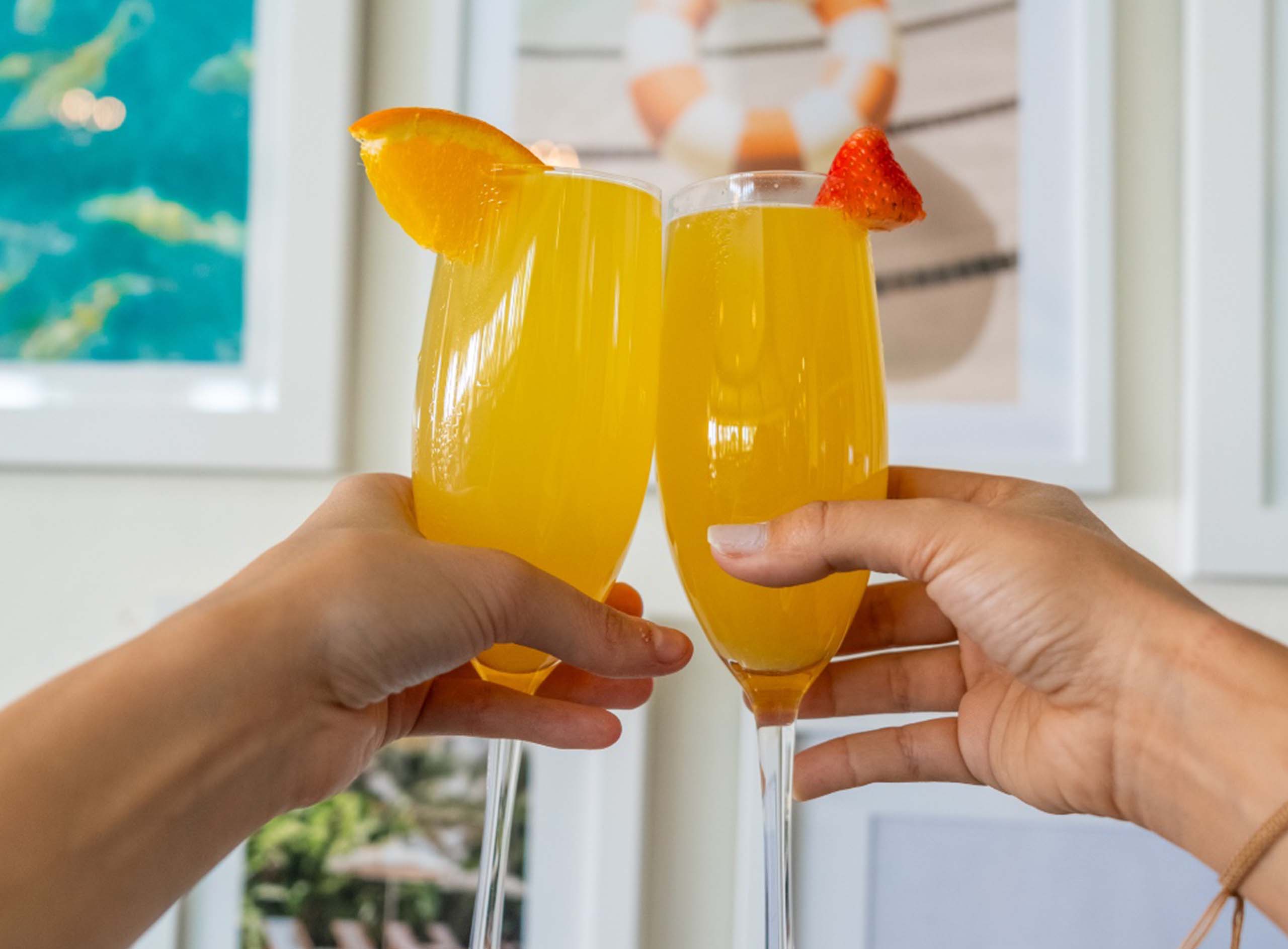 two girls toasting with two mimosas with a fruit garnish on each